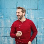 Home A joyful man in a red sweater smiles while listening to music on his smartphone against a blue brick wall.